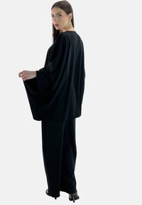 Schwarzes Oberteil mit weiten Ärmeln, kombiniert mit lockeren schwarzen Hosen aus glattem Stoff, die ein entspanntes Design und subtile Drapierungsdetails aufweisen.