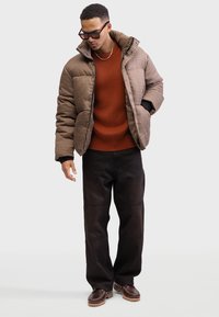 Ruskea houndstooth-tyylinen puuhkatakki mustilla yksityiskohdilla, päällä rustonpunainen ribbipaita. Yhdistetty tummiin housuihin ja ruskeisiin jalkineisiin.