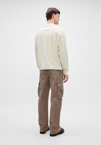 Ανοιχτό μπεζ φούτερ με στρογγυλή λαιμόκοψη, συνδυασμένο με χαλαρές καρό παντελόνες σε κιτρινωπό καφέ με πλαϊνές τσέπες και ίσια γραμμή.