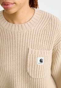 Maglione beige lavorato a maglia con texture a coste, scollo rotondo e una tasca frontale che presenta un dettaglio con logo in bianco e nero.