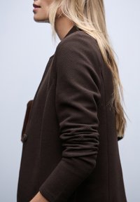 Hnědý strukturovaný blazer s uvolněným střihem, disponující jedním knoflíkem a ohrnutými rukávy, zvýrazněný na světlém pozadí.