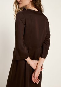 Frau mit lockigem Haar, die ein langärmliges, dunkelbraunes Kleid trägt, steht mit gefalteten Händen hinter ihrem Rücken und schaut seitlich.