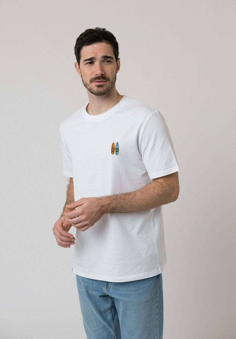 Man in white t-shirt with small orange and blue surfboard prints on chest, wearing light blue jeans, standing against plain light background.