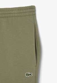 Olijfgroene joggingbroek van zacht materiaal, met een zijzak en een klein geborduurd krokodillenlogo in de rechterbenedenhoek.
