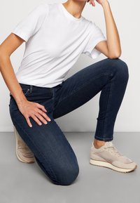 T-shirt bianca in cotone, jeans skinny blu scuro e sneakers beige con una tomaia testurizzata. L'individuo è in ginocchio, con la mano appoggiata sul ginocchio.