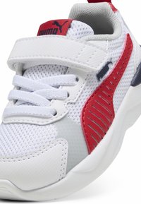 Baskets blanches de sport avec dessus en maille, accents rouges et gris, bande Velcro et semelle texturée pour l'adhérence. Logo Puma sur la languette.