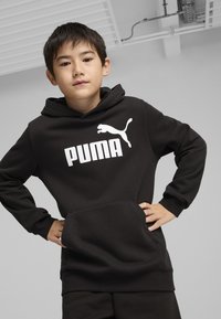 Crna dukserica s kapuljačom i kengur džepon, s bijelim "PUMA" logotipom i siluetom skakačkog pumina. Mekani materijal, ležeran kroj.