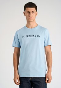 T-shirt di cotone azzurro chiaro con maniche corte, con la scritta "COPENHAGEN" in stampa scura. Scollo rotondo e tessuto liscio, abbinata a jeans scuri.