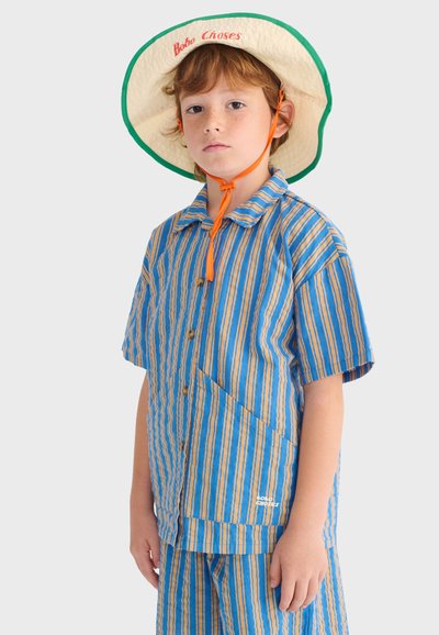 Enfant à la peau claire et aux cheveux roux portant un chapeau seau crème avec une bordure verte et un cordon orange, ainsi qu'un t-shirt à manches courtes rayé bleu et orange et un short assorti.