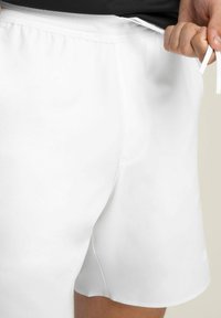 Bermudas deportivas blancas con una cinturilla texturizada y cordón. Tela suave, sin patrones visibles y una ligera abertura lateral en el dobladillo.