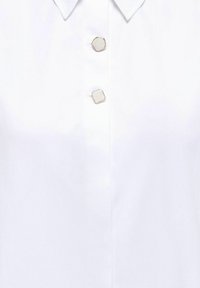 Chemise à col blanc avec deux boutons carrés irréguliers de couleur neutre claire, placés au centre de la patte de boutonnage avant.