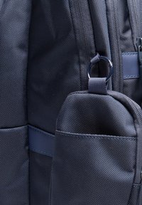 Tumesinise nailonist seljakott, millel on tekstuuriga kangas, mitmed tõmblukud ja küljetasku väikese silmusega aksessuaaride kinnitamiseks.