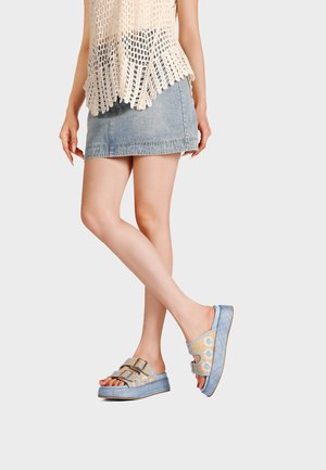 Gambe incrociate che indossano una minigonna di denim blu e sandali platform con punta aperta, fibbie e cinturini beige con motivo floreale.