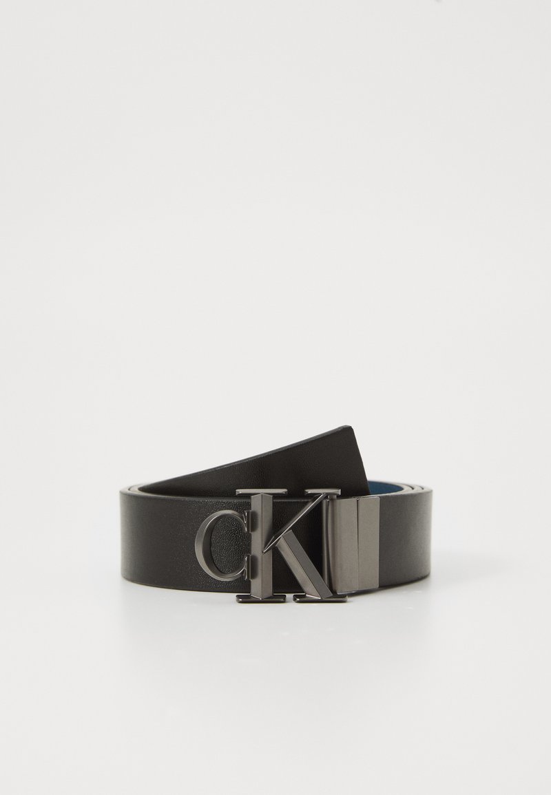 Zwarte leren riem opgerold met glanzende zilveren "CK" gesp op een witte achtergrond.