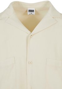 Chemise à manches courtes couleur crème en tissu lisse, avec un col en V, deux poches avant et une fermeture à boutons.