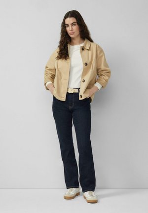 Jeune femme aux longs cheveux bouclés portant une veste beige boutonnée, un haut blanc, un jean foncé, des baskets blanches et une ceinture, debout les mains dans les poches.