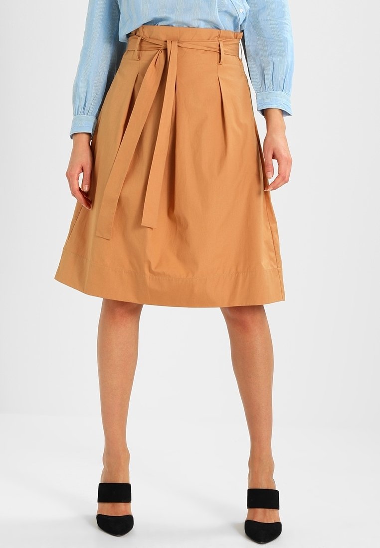 Femme portant une jupe beige longueur genoux avec une ceinture nouée, une chemise bleu clair à manches longues et des talons noirs à brides, debout devant un fond uni.