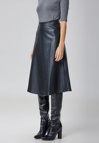 Jupe en cuir noir en forme de A avec une taille haute, texture lisse et design longueur genou, associée à des bottes hautes vernies noires à motif crocodile.