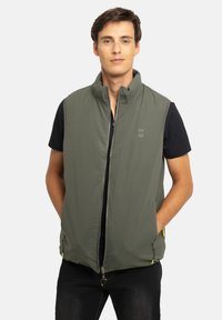 Veste sans manches vert olive avec col montant, fermeture éclair et poches latérales. Comprend des détails subtils du logo et des accents jaunes contrastants.