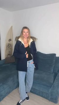 Casaco azul-marinho com capuz forrado a pelo, combinado com um top preto e jeans largos azul-claro, em pé em um sofá azul com uma prancha de surf próxima.