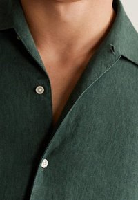 Camisa verde de lino con cuello y dos botones, con un tejido texturizado y un brillo sutil. Vista en primer plano del escote y los botones.