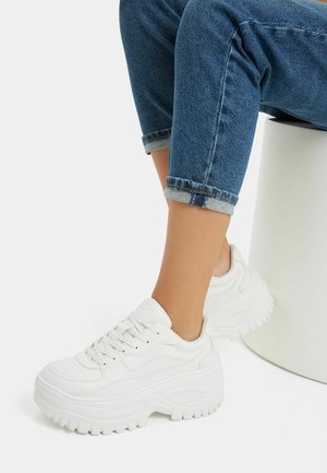 Persoon die opgerolde blauwe jeans en dikke witte plateau-sneakers draagt, zittend op een wit cilindrisch krukje tegen een effen achtergrond.