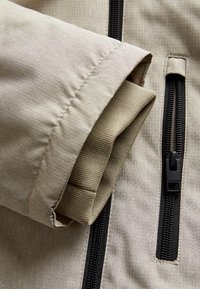 Manche de veste beige avec un tissu texturé, présentant un poignet replié contrastant et deux zippers noirs pour des accents fonctionnels.
