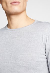 Hellgraues langärmliges Baumwollshirt mit Rundhalsausschnitt. Sichtbare Strickstruktur, glatte Oberfläche und minimale Nähdetails.