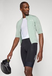 Maillot de cyclisme vert clair à manches courtes avec zip, associé à un short de cyclisme rembourré noir avec un logo et des accents blancs. Casque noir tenu à la main.
