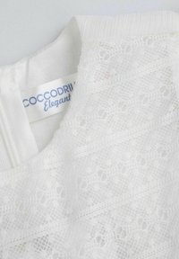 Biała koronkowa bluzka z delikatnym wzorem i kwiatowymi akcentami, gładkim białym wykończeniem oraz metką z napisem "Cocodrillo Elegant."