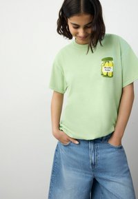 T-shirt à manches courtes vert clair avec un pot de cornichons imprimé et le texte "Pleasantly Pickled" sur la poitrine gauche. Matière en coton doux.