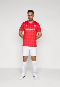 Camisola de futebol vermelha com padrão de losangos, apresentando logótipo e texto brancos. Combinada com calções e meias brancas, as meias têm padrões claros.