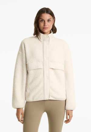 Vrouw in een crèmekleurige fleecejack met drukknoopjes en twee voorzakken, gecombineerd met beige textuurleggings, op een witte achtergrond.