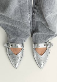 Ballerine metalliche argento a punta con fianchi a taglio, dotate di borchie tonde e fibbia laterale. Indossate con jeans grigi chiari e larghi.
