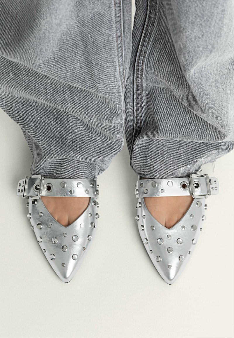 Ballerine metalliche argento a punta con fianchi a taglio, dotate di borchie tonde e fibbia laterale. Indossate con jeans grigi chiari e larghi.
