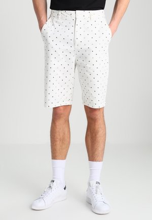 Mężczyzna ubrany w białe szorty z małymi czarnymi geometrycznymi wzorami, białe skarpetki i białe tenisówki, stoi z rękami w kieszeniach.