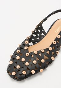 Sandal nera in sintetico caratterizzata da un design con ritagli e borchie metalliche dorate, con punta aperta, cinturino slingback e una soletta beige liscia.