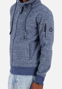Mężczyzna w niebieskiej, melanżowej bluzie z kapturem, z kieszeniami z przodu i zapinaną na zamek kieszenią na lewym rękawie, w połączeniu z jasnoniebieskimi dżinsami.