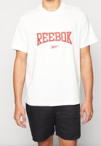 Vit bomull t-shirt med kort ärm, med en framträdande röd "REEBOK" textlogotyp och en liten röd grafik under, i kombination med svarta shorts.