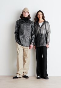 Deux individus portent des vestes de pluie légères et transparentes de couleur grise, avec des boutons et des poches, associées à des pantalons beiges et noirs et à des chaussures décontractées.