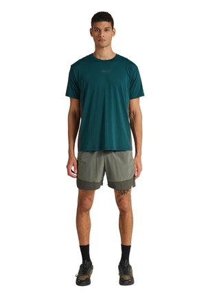 Mand stående med fronten mod kamera iført en mørkegrøn kortærmet skjorte, olivengrønne shorts, sorte sokker og sorte sneakers på hvid baggrund.