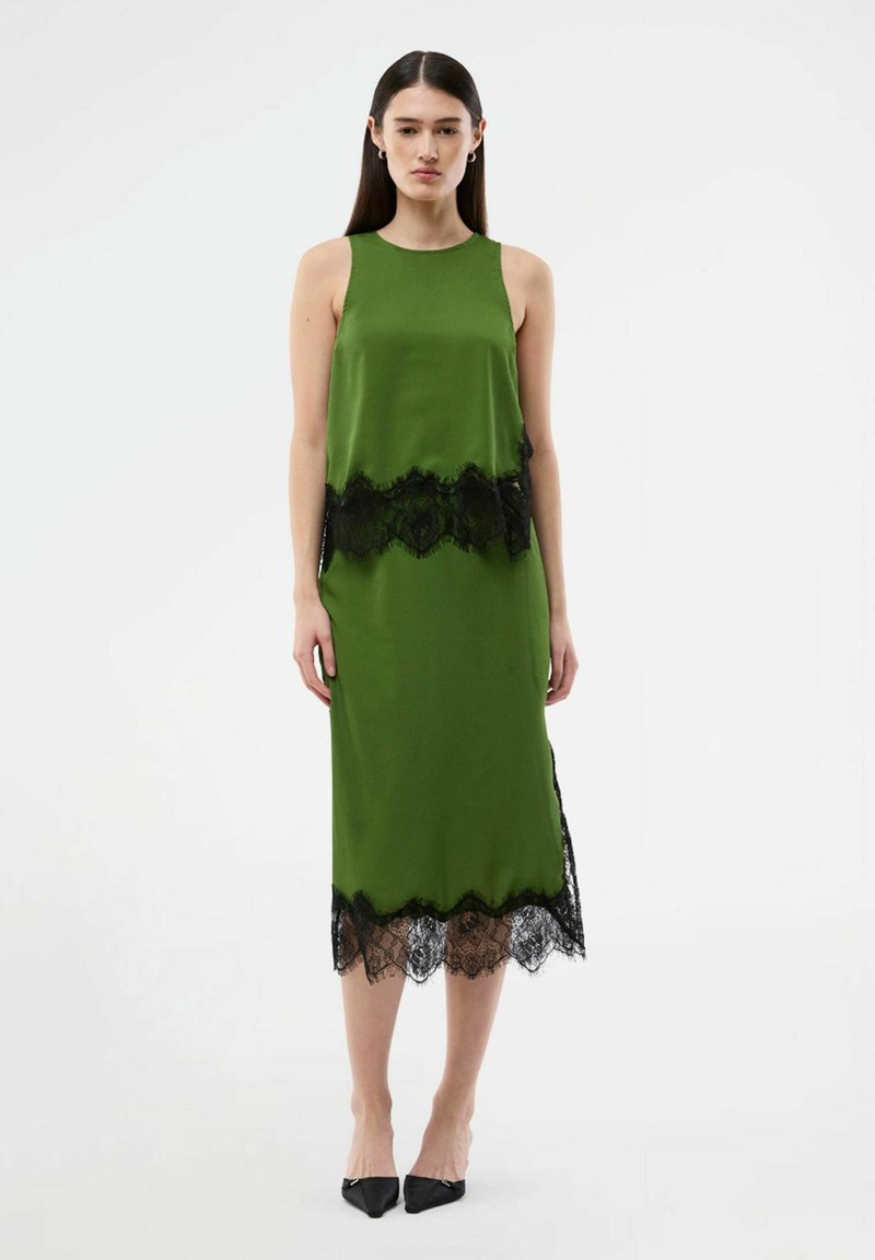 Femme debout portant une robe midi verte sans manches avec une bordure en dentelle noire à la taille et à l'ourlet, associée à des talons pointus noirs.