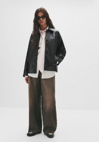 Schwarze Lederjacke mit Knopfverschluss, kombiniert mit einem hellfarbenen Hemd und weiten braunen Denim-Jeans. Schwarze Sonnenbrille rundet den Look ab.