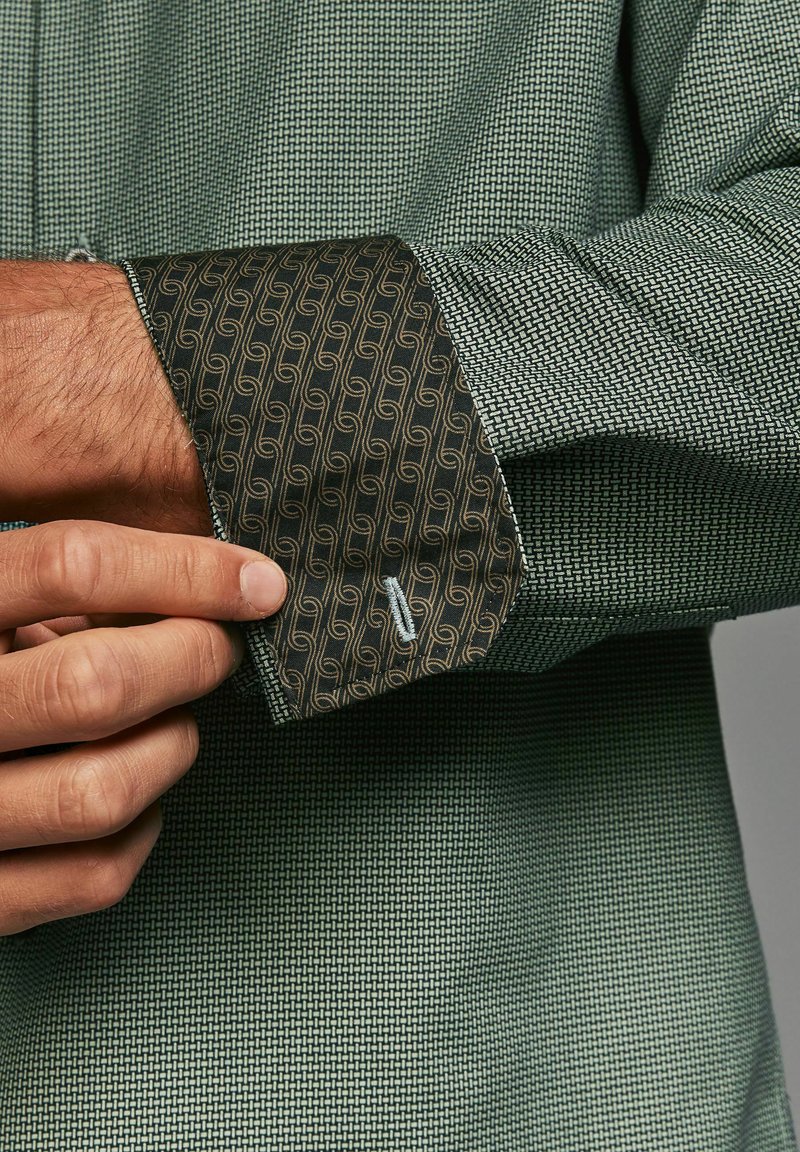 Green patterned shirt featuring a textured weave. Cuffs display a dark fabric with a gold swirl pattern and contrasting stitching detail.