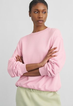 Žena v světle růžové mikině s vyhrnutými rukávy a světle zelených kalhotách, stojící se založenýma rukama a dívá se stranou.