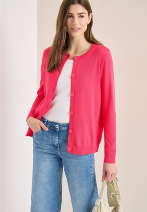 Donna che indossa un cardigan rosa acceso con bottoni sopra una maglietta bianca e jeans blu, che tiene una borsa beige in una mano.