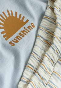 Tissu bleu clair avec un graphique demi-soleil marron et le texte « sunshine » à côté d’un tissu blanc avec des rayures bleues et marron et une texture froncée.