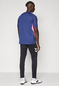 Moški z na spletenimi lasmi obrnjen stran, oblečen v modro-roza športno majico, črne športne hlače z belimi chevron logotipi in bele športne čevlje.