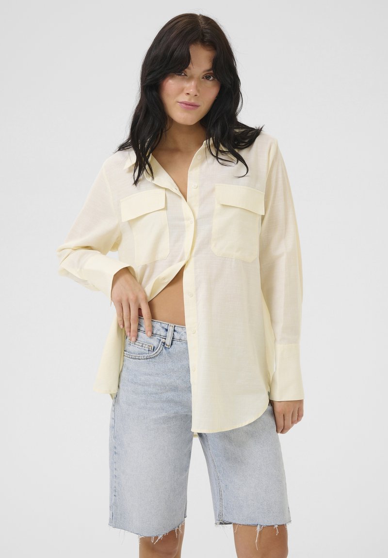 Chemise à manches longues, jaune clair, avec boutons sur le devant, deux poches poitrine, et coupe décontractée, associée à un short en denim usé.