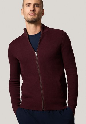 Mężczyzna w burgundowym, strukturalnym swetrze na zamek, noszącym granatową koszulę, z rękami w kieszeniach ciemnych spodni, stojący na tle jasnego, jednolitego tła.